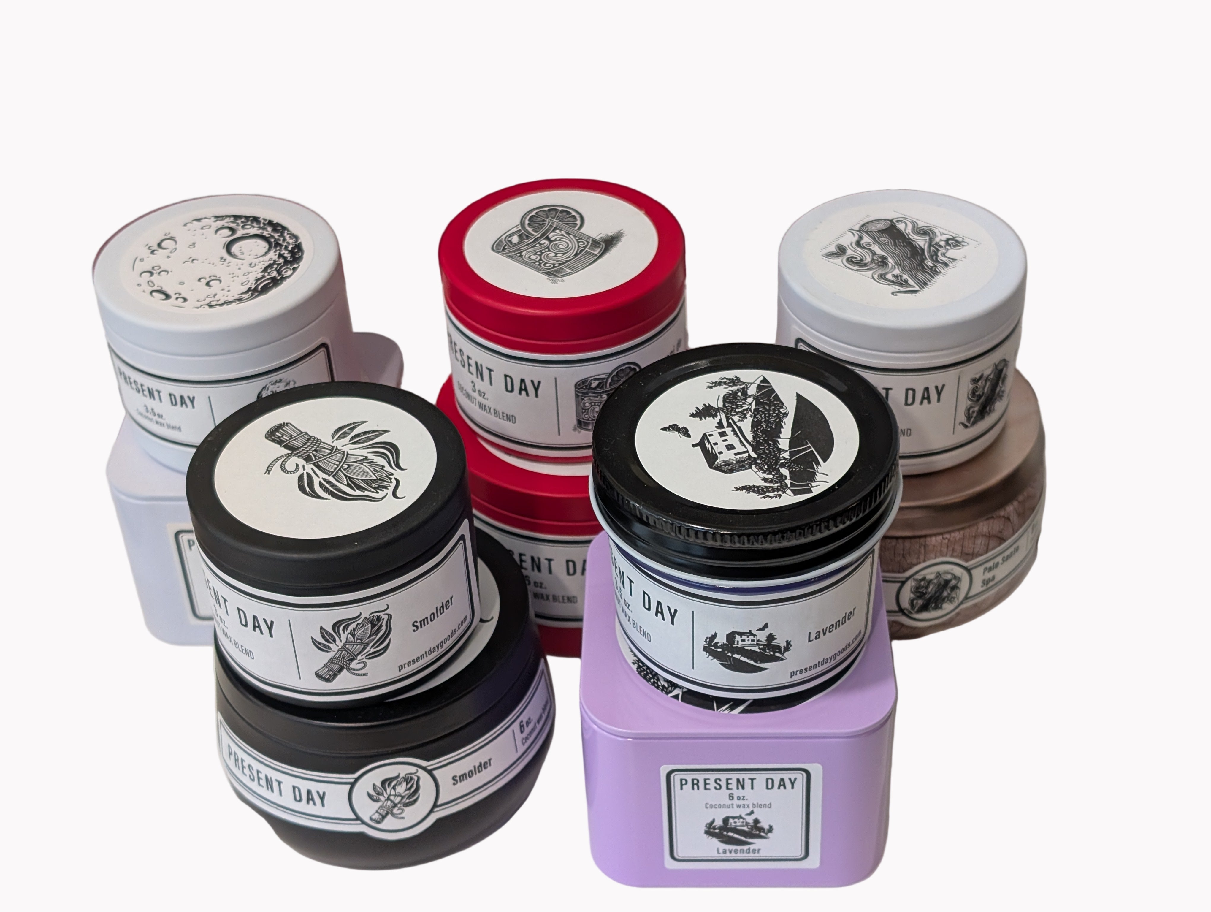Collection of 'Present Day' product tins on a surface with a smolder, lavender, palo santo, negroni de mare and Moonflower and pear candle setup.