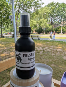 Black spray bottle labeled 'Present Day Goods' on a wooden surface with a park in the background
