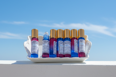 Six small cosmetic roller bottles with gold caps and blue, purple, and red liquids, displayed in a white ceramic tray on a white ledge against a bright blue sky.
