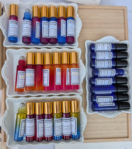 An overhead display of multiple essential oil roller bottles in assorted colors, arranged in white trays on a wooden board. The bottles feature gold and black caps, with white labels showing the brand name 'PRESENT'.