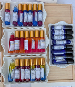 An overhead display of multiple essential oil roller bottles in assorted colors, arranged in white trays on a wooden board. The bottles feature gold and black caps, with white labels showing the brand name 'PRESENT'.