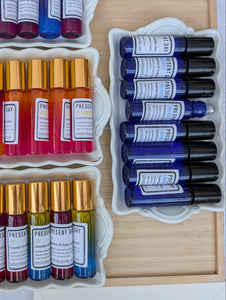 An overhead display of multiple essential oil roller bottles in assorted colors, arranged in white trays on a wooden board. The bottles feature gold and black caps, with white labels showing the brand name 'PRESENT'.