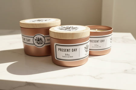 Three 'Present Day' candles with wooden tin printed lids on a light surface.