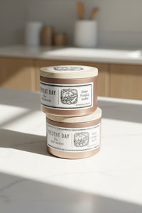 Two 'Present Day' candles stacked with a bowl of oranges in the background