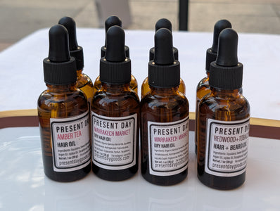 Six amber glass dropper bottles of hair and beard oils arranged on a white surface. The bottles have black droppers and labels for Present Day Hair & Beard Products, including Amber Tea, Marrakesh Marmalade, and Redwood + Tobacco.