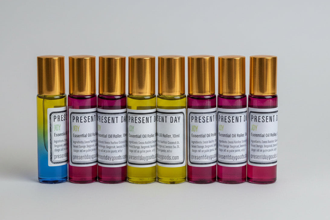 Six essential oil roller bottles with gold caps arranged in a white ceramic tray, alongside a blue-and-yellow gradient roller bottle. The labels read 'PRESENT DAY' and 'Essential Oil Roller Bottles'.