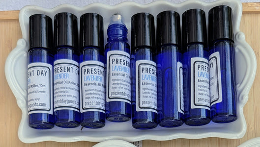 A set of essential oil roller bottles in cobalt blue glass with black caps, arranged in a white ceramic tray. The labels show different scents including Sandalwood, Lavender, and Enzyme.