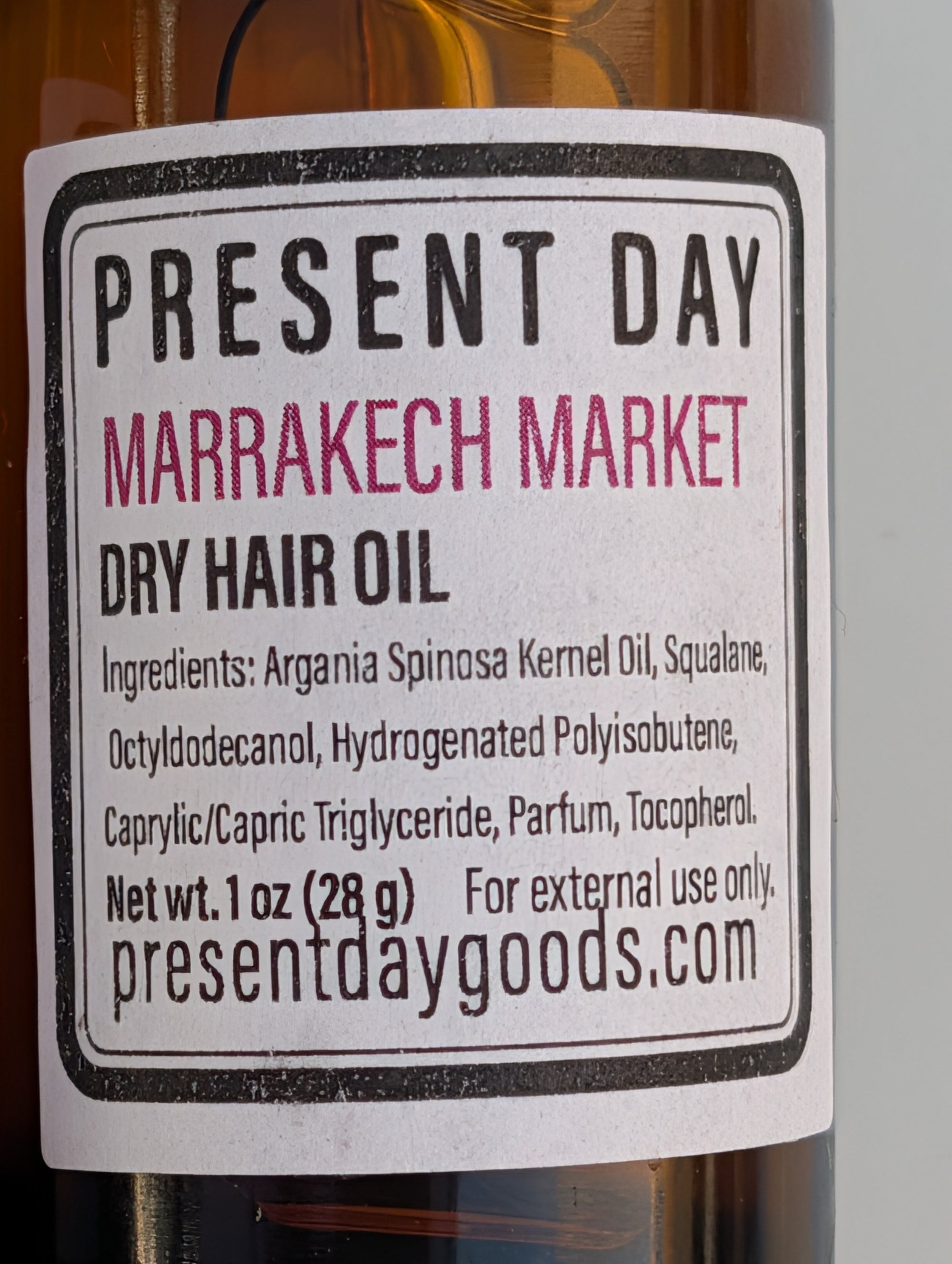 A close-up of a bottle label for Present Day Marrakech Market Dry Hair Oil, showing amber liquid in a glass bottle with a white label and black text.