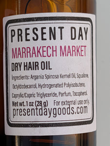 A close-up of a bottle label for Present Day Marrakech Market Dry Hair Oil, showing amber liquid in a glass bottle with a white label and black text.