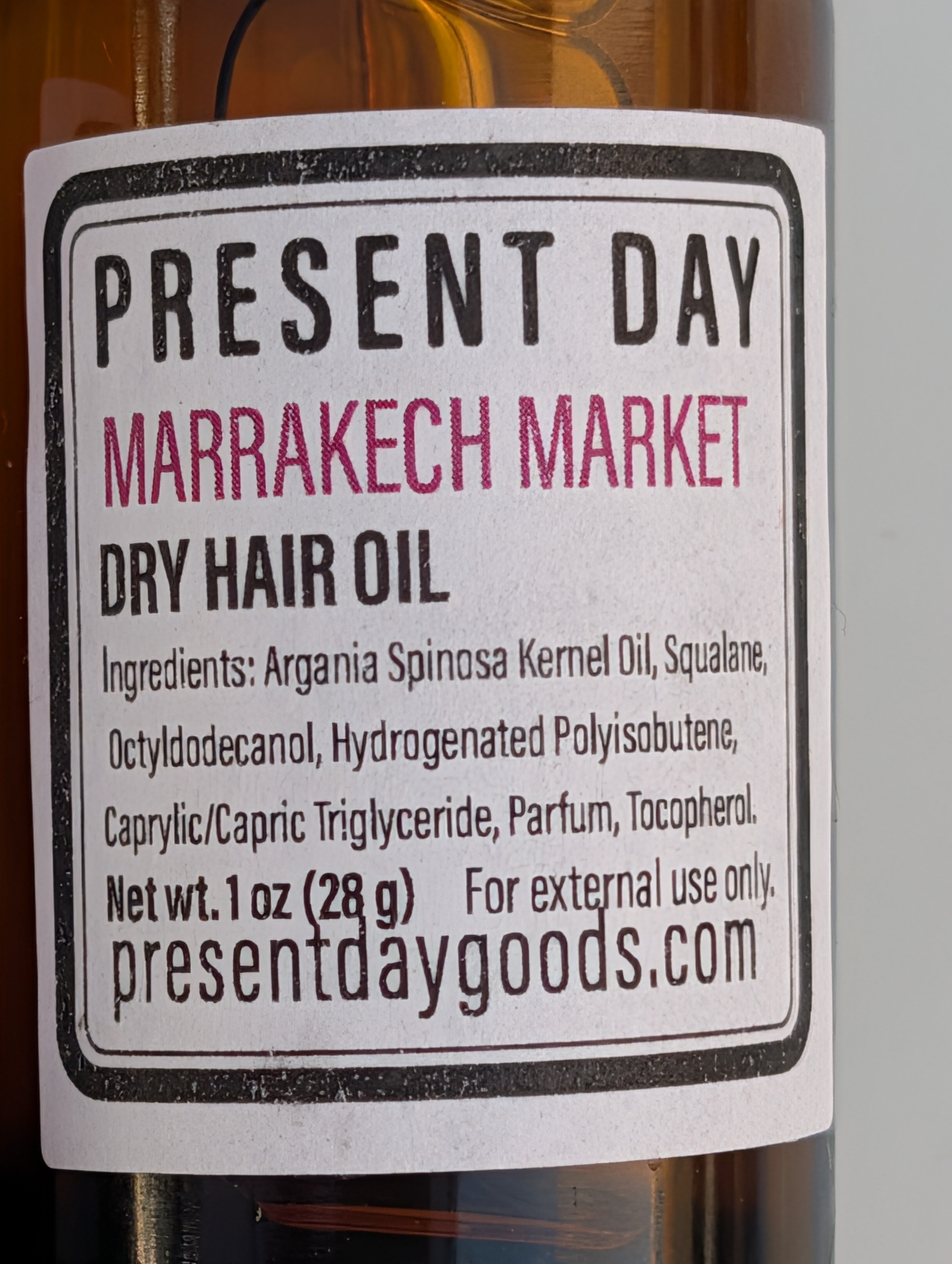 A close-up of a bottle label for Present Day Marrakech Market Dry Hair Oil, showing amber liquid in a glass bottle with a white label and black text.