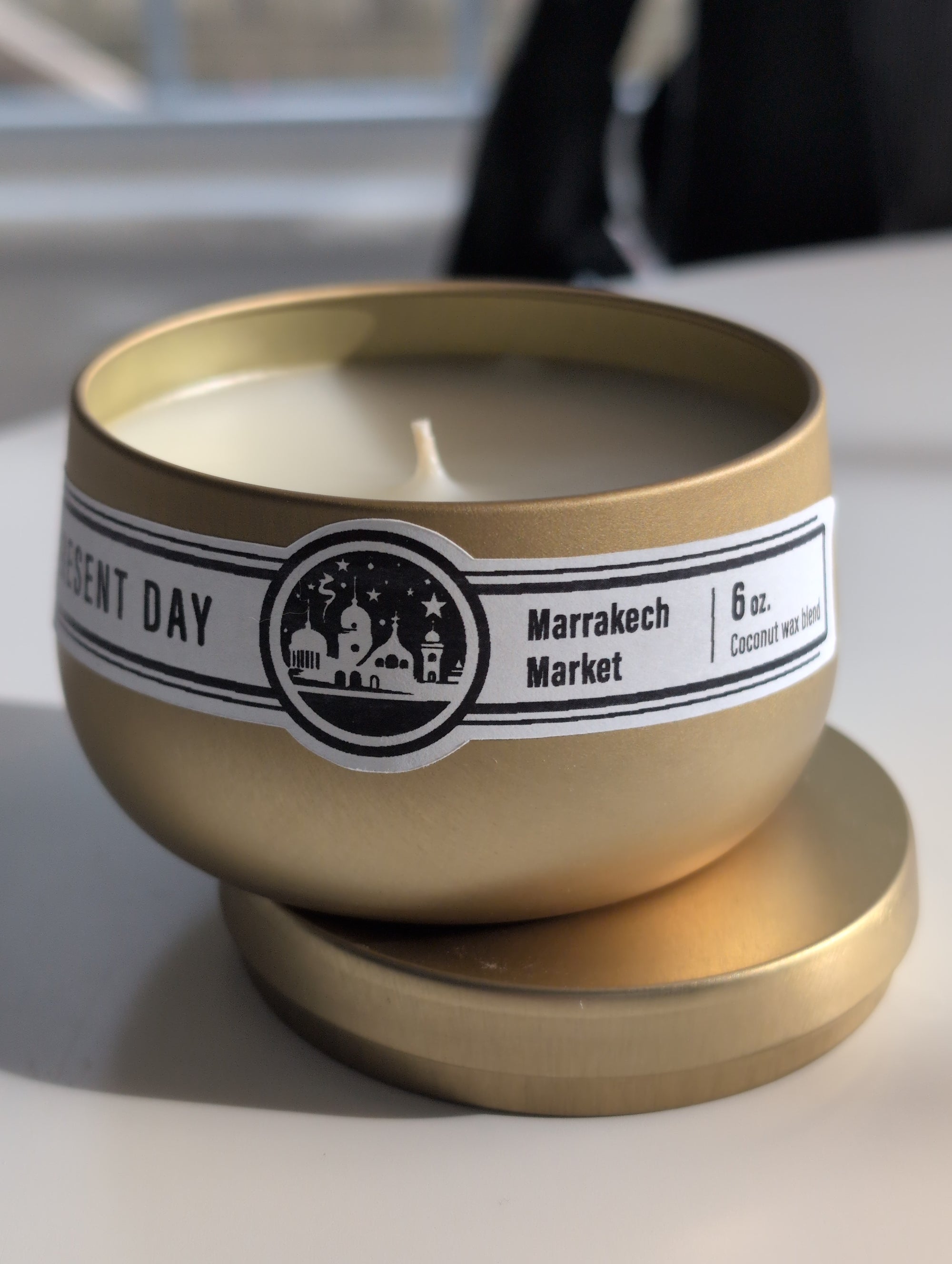 A gold-colored metal tin lid placed on top of a matching tin, which contains a scented candle. The label on the tin reads 'Marrakech Market 6 oz.