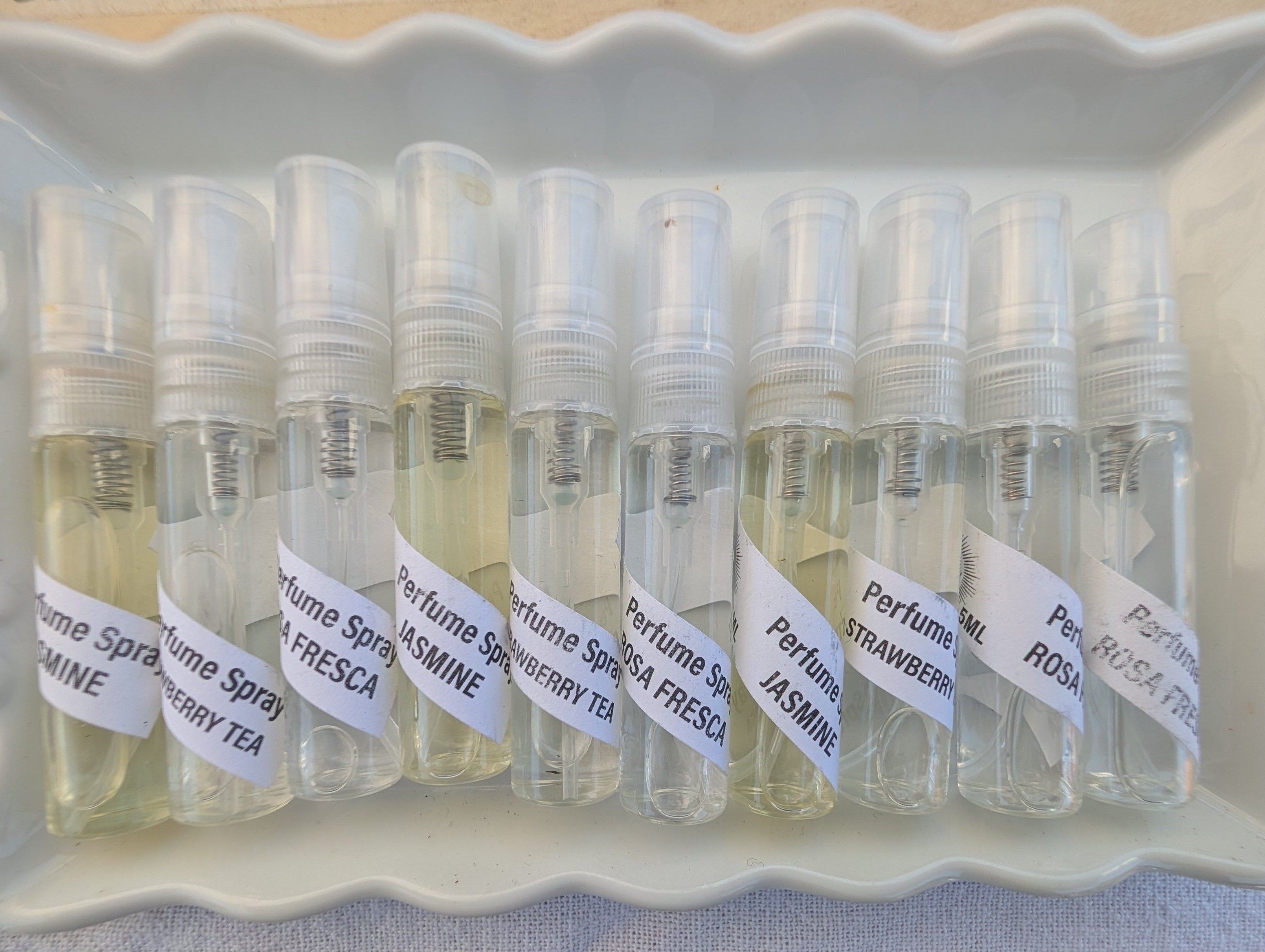 A tray of small clear glass perfume sample vials with white labels, photographed from above on a light surface next to a printed box.