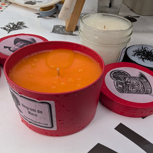 Two candles in red containers with labels on a white surface with black designs.