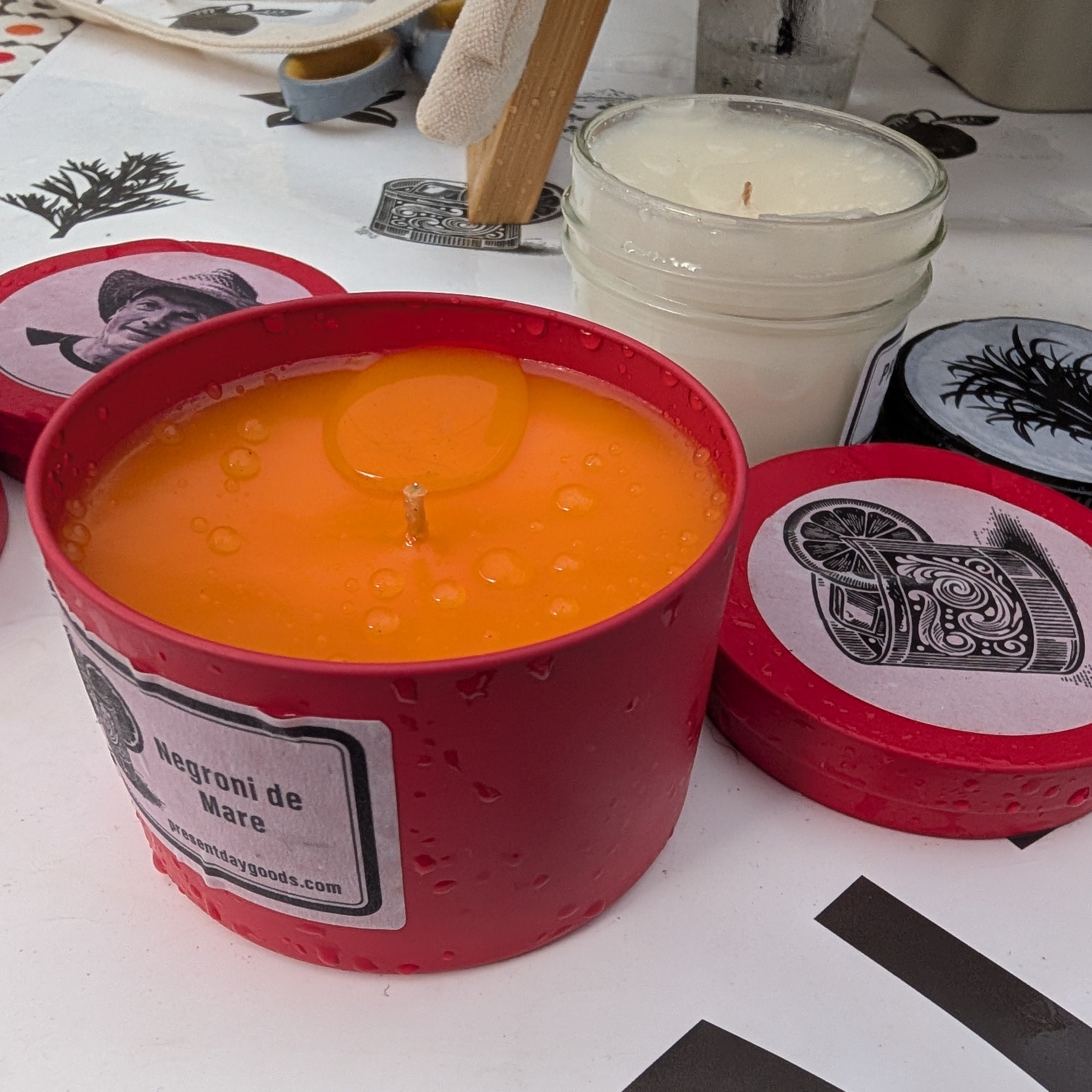 Two candles in red containers with labels on a white surface with black designs.