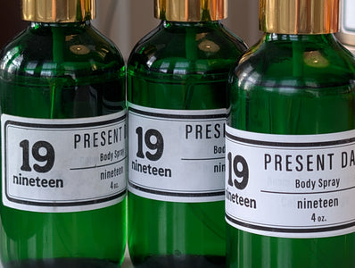 Three bottles of 'Present Day' body spray with gold caps and white labels.