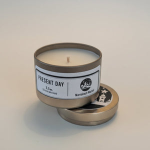 Candle in a metal tin with 'Present Day' label on a light background