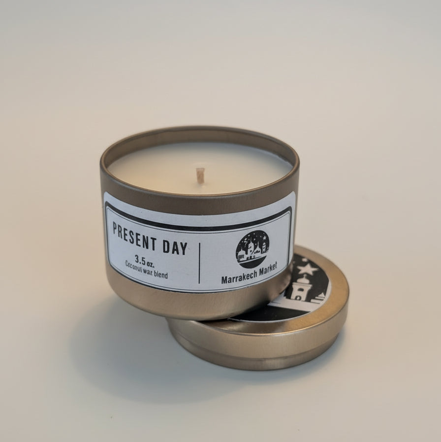 Candle in a metal tin with 'Present Day' label on a light background