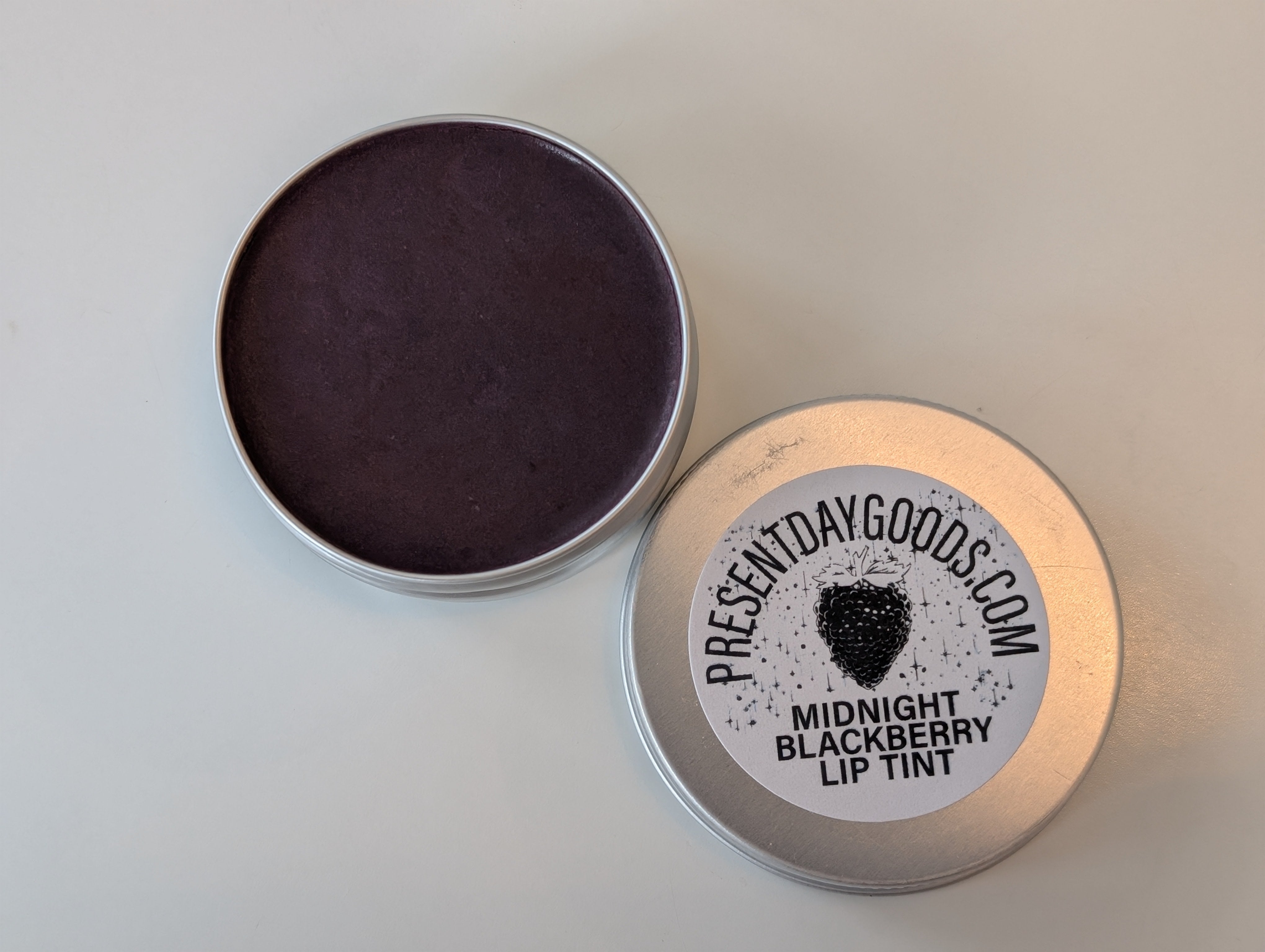 Tin of Midnight Blackberry lip tint with 'Present Day Goods' label on a light gray background