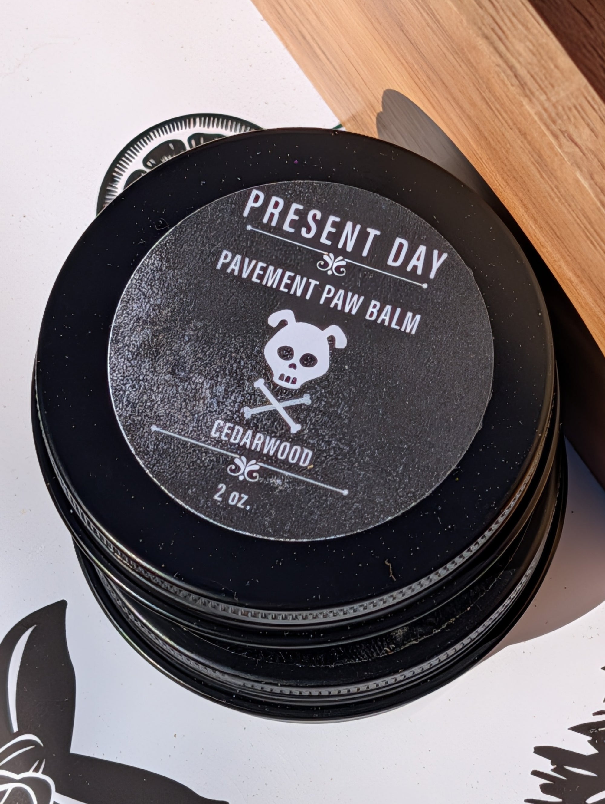 Stack of 'Present Day' pavement paw balm containers with a wooden board in the background.