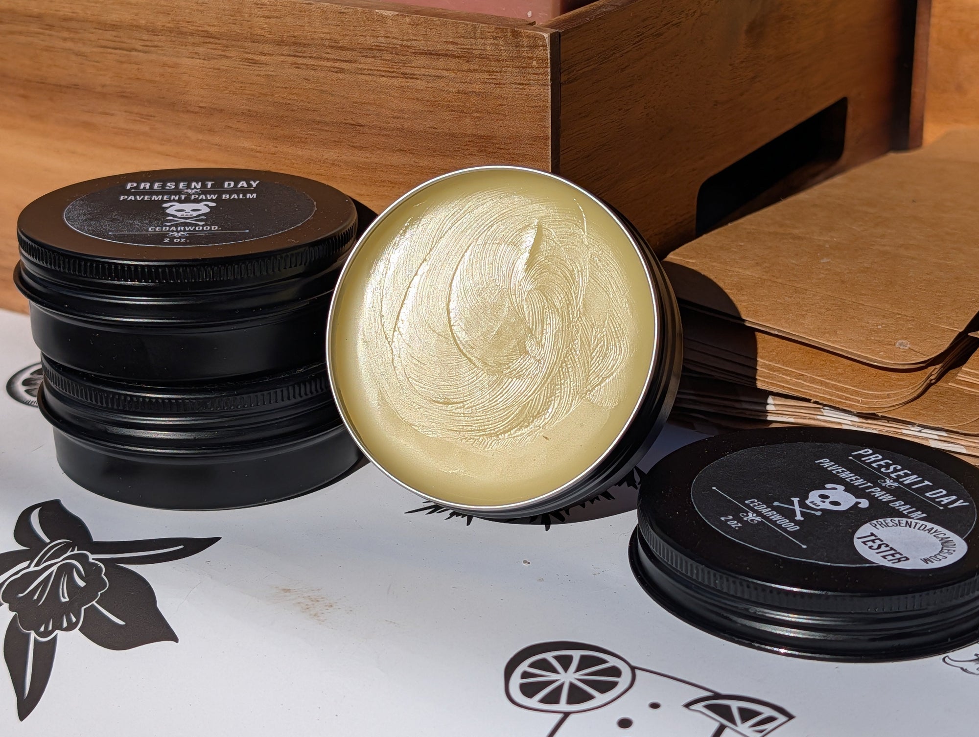 Open container of cream with black lids on a branded paper, with wooden shelves in the background.