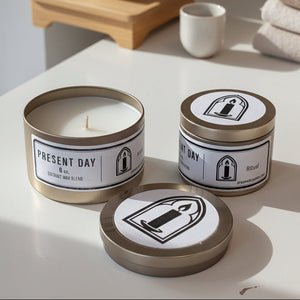 Two candles in tins with 'Present Day' labels on a white surface.