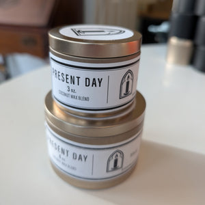 Two 'Present Day' candles stacked on a surface with a blurred background