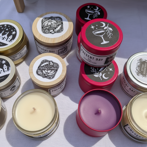 Collection of candles in various tins with decorative labels on a light surface.