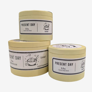 Three 'Present Day' candles on a white surface with a blurred background