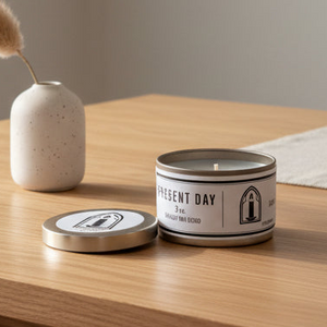 Candle in a tin with a label on a wooden surface next to a vase.