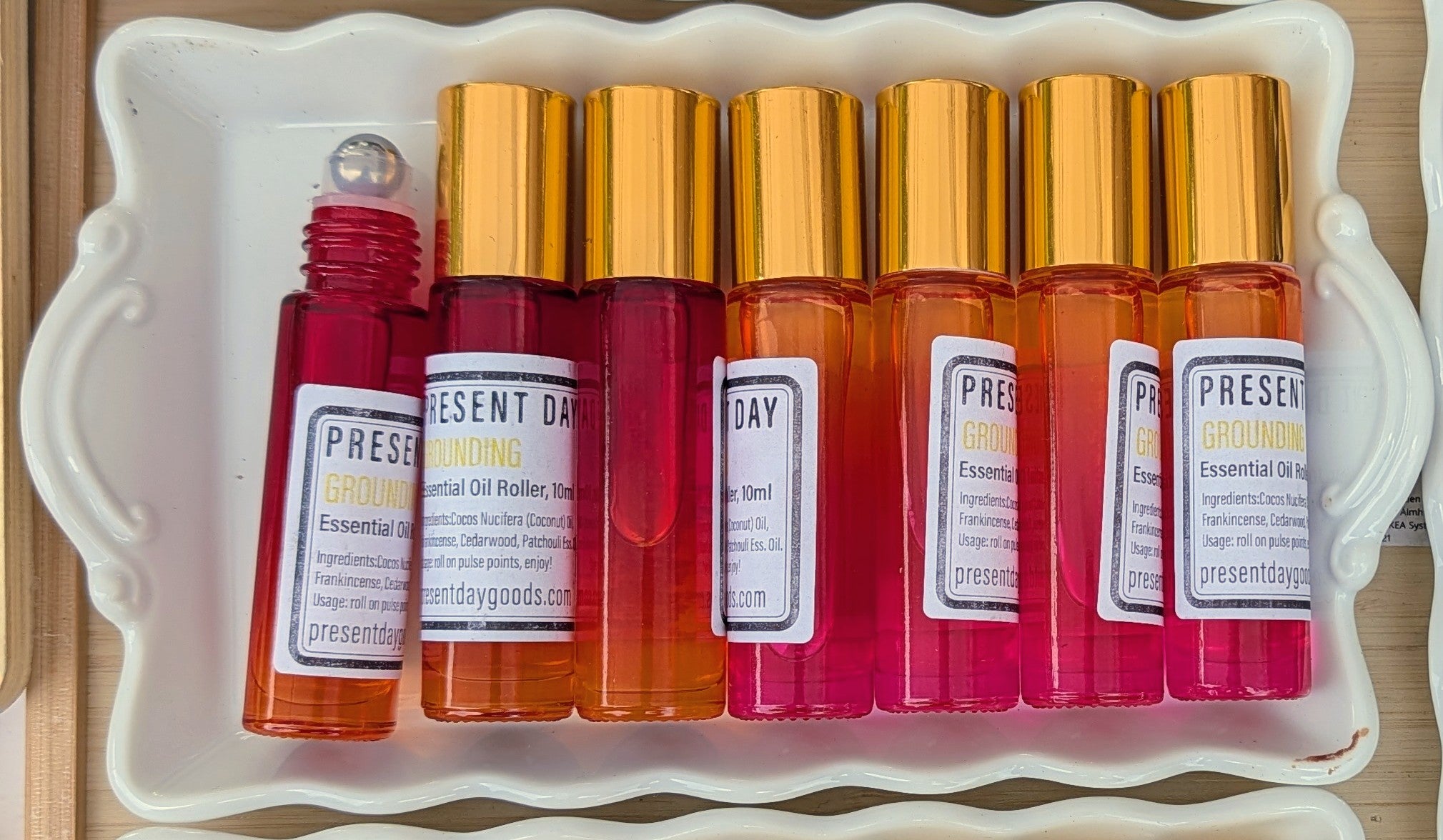 Six small essential oil roller bottles with gold caps and white labels arranged in a white ceramic tray. The bottles contain orange, pink, and red liquids, with the leftmost bottle uncapped to show the roller ball and liquid.
