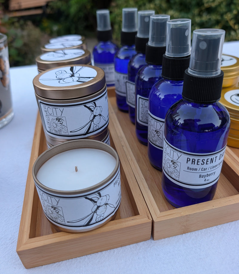 A collection of scented candles and spray mists from the brand 'Present Day Candles' arranged on bamboo trays outdoors on a white tablecloth, with green foliage blurred in the background.