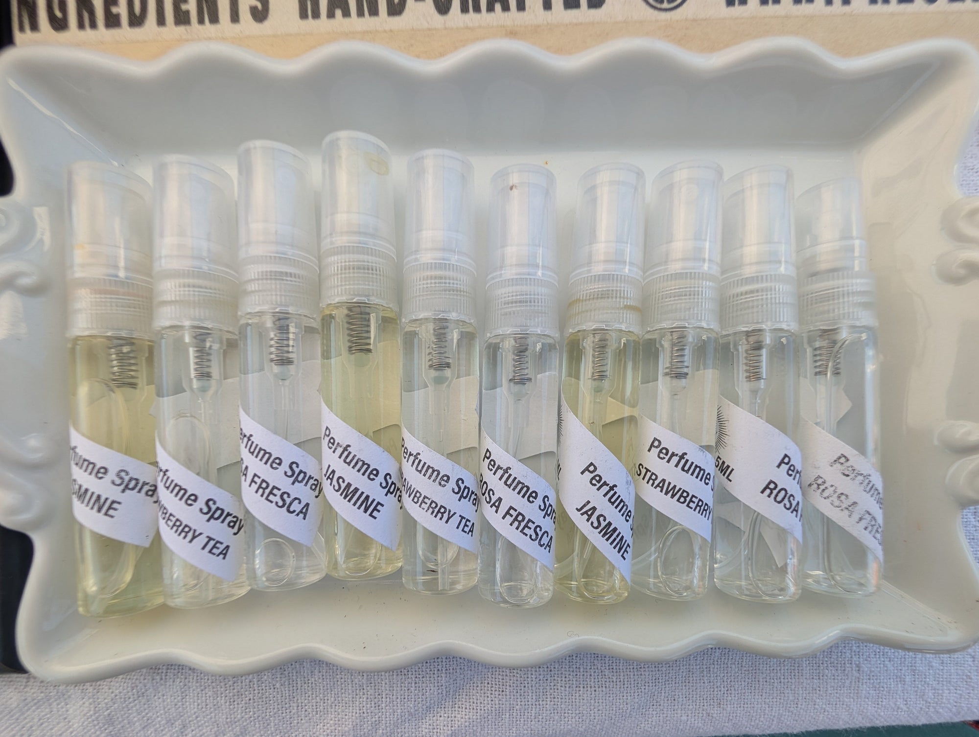 A tray of small clear glass perfume sample vials with white labels, photographed from above on a light surface next to a printed box.