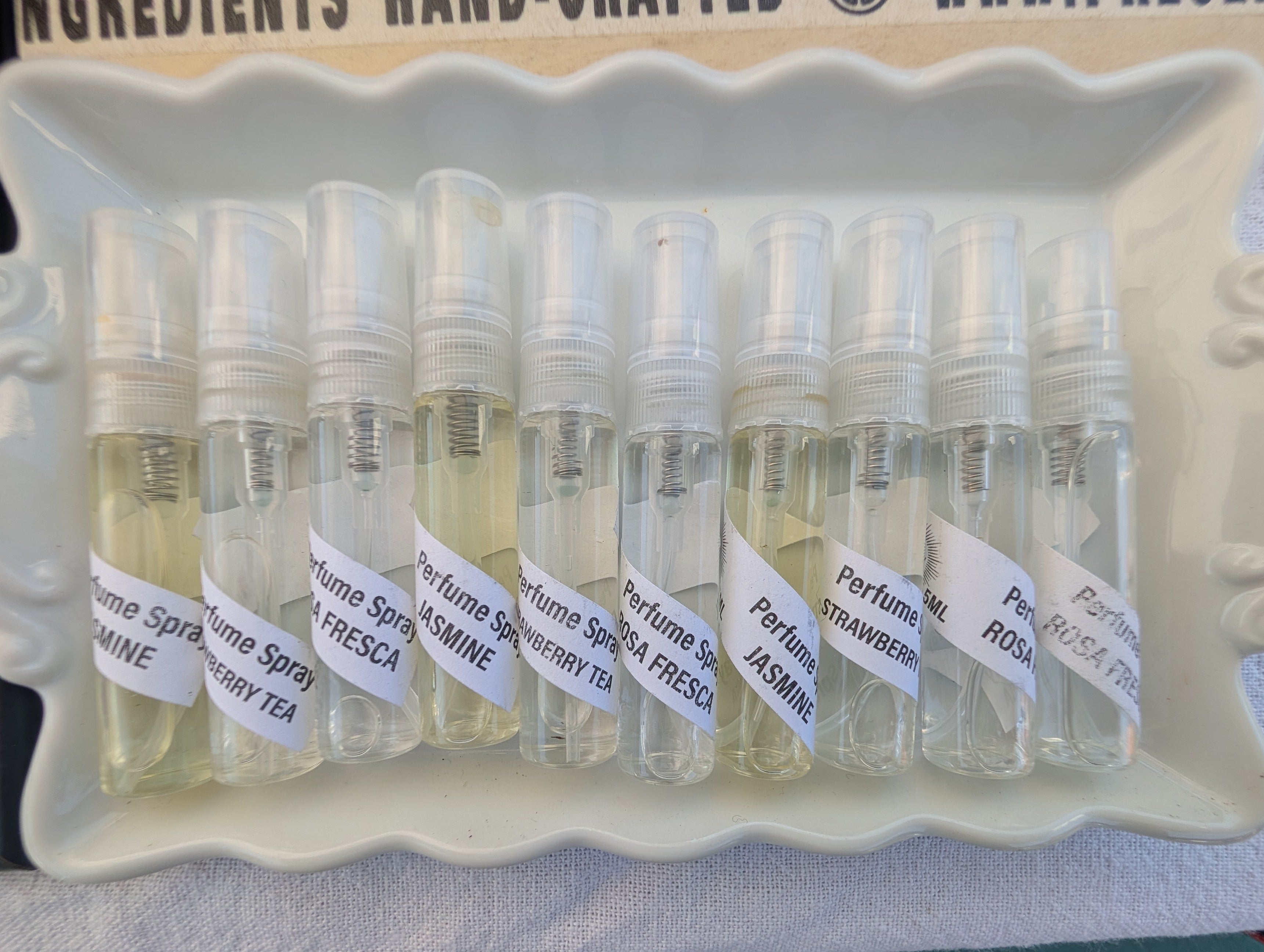 A tray of small clear glass perfume sample vials with white labels, photographed from above on a light surface next to a printed box.