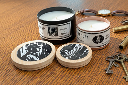Two candles with labels and wooden print tin lids on a wooden surface 