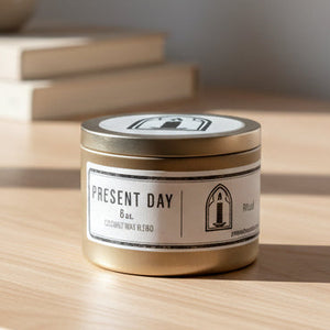 Candle in a metal tin labeled 'Present Day' on a white surface.