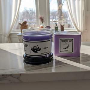 Two candles with 'Present Day' branding on a marble surface in front of a window.